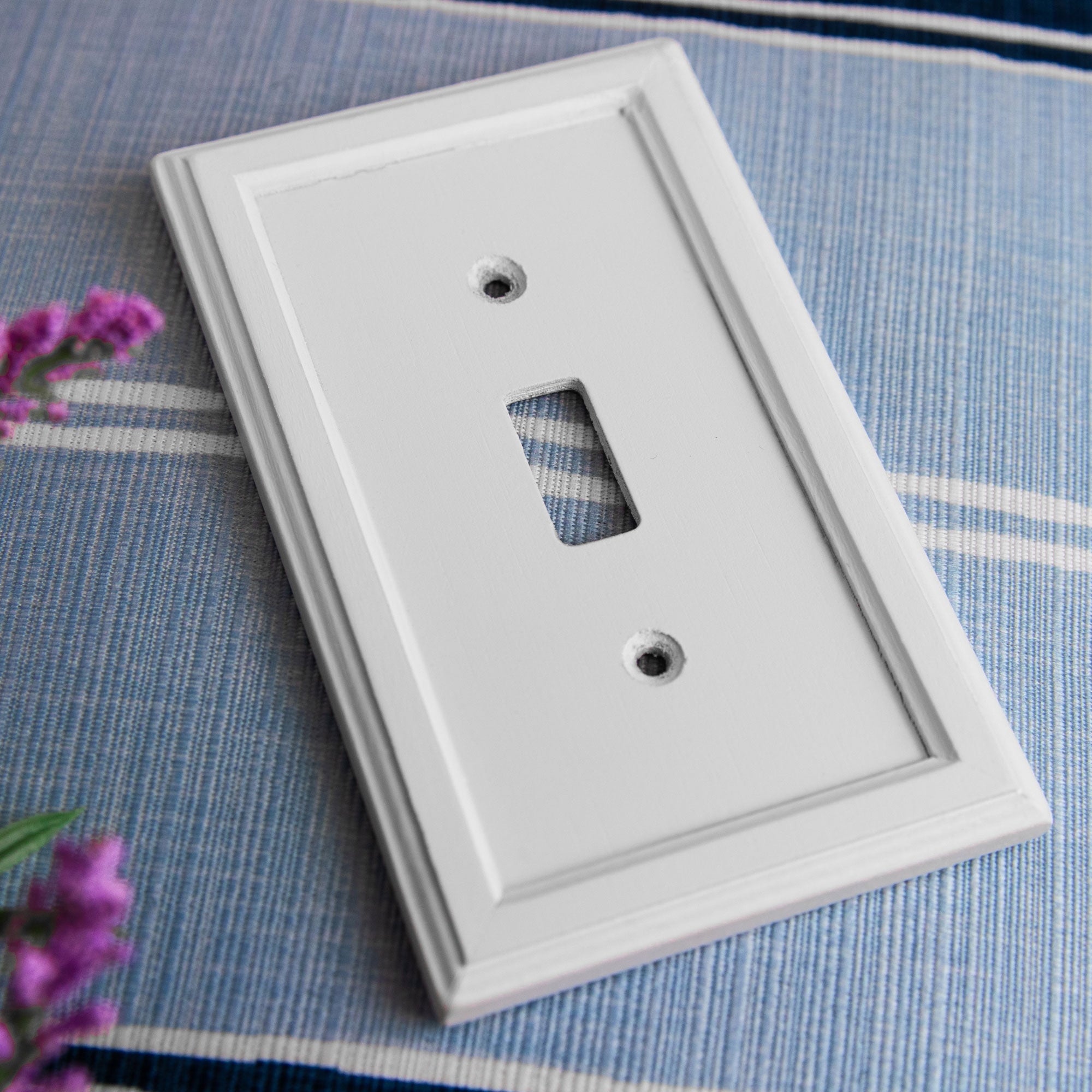 A white light switch cover from the Elly collection, showcasing a smooth, matte finish with clean, minimalist lines that enhance its classic charm. The crisp white surface offers a fresh and timeless look, seamlessly blending with any decor. It is lying on a soft blue blanket.