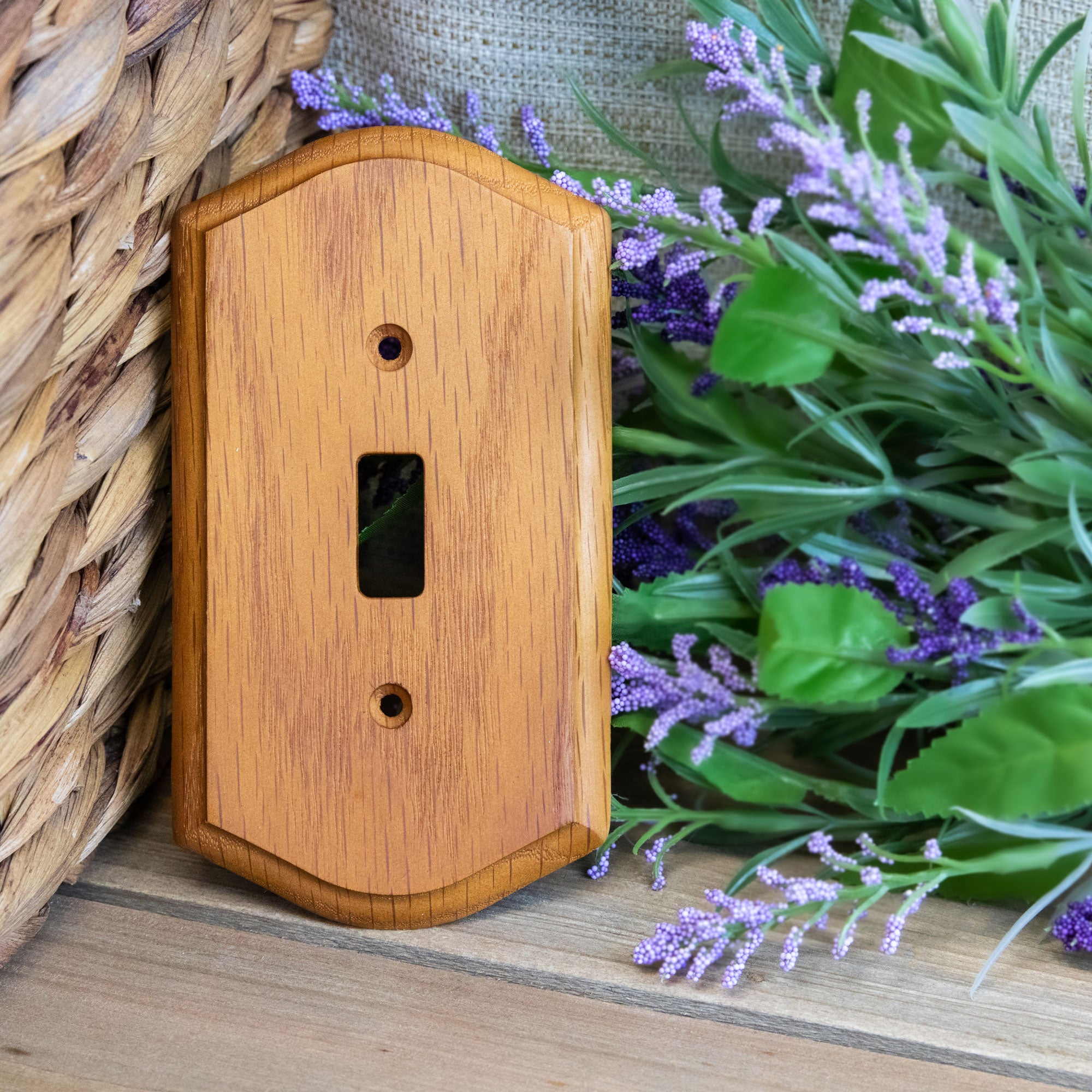 
Country-style medium oak light switch wallplate with a warm, natural wood grain and slightly weathered finish. The wallplate is propped up again a woven basket next to a lavender plant.